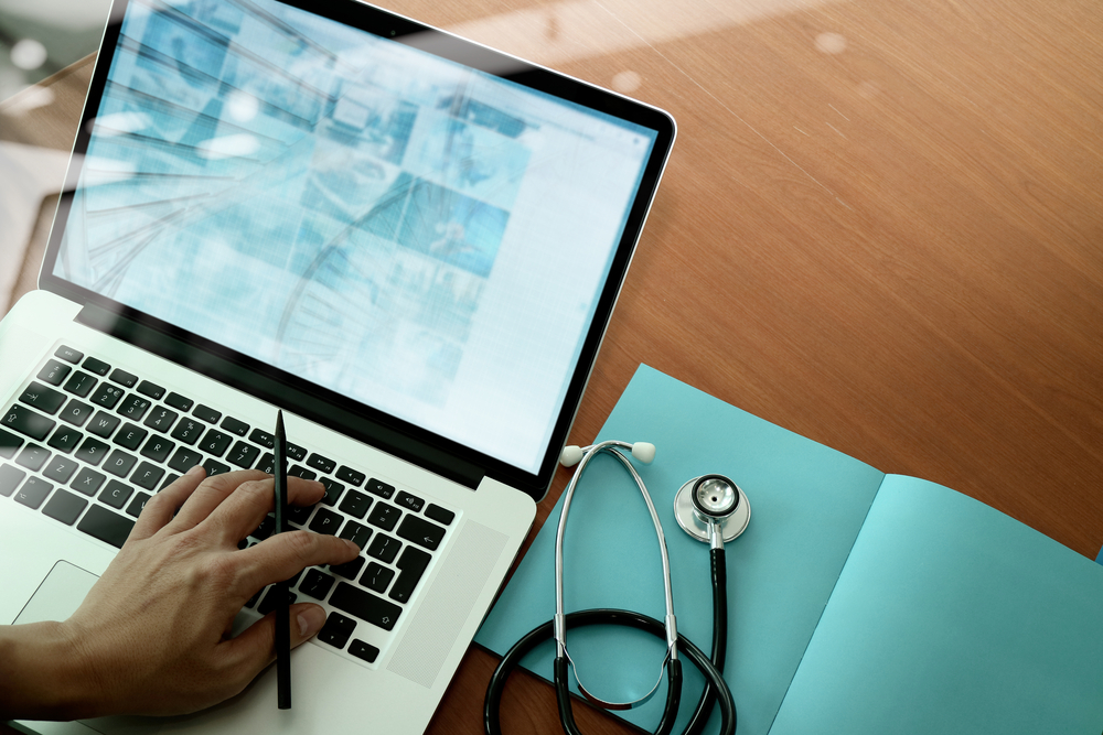 close up hand holding a pen above laptop keyboard next to notepad and stethoscope 